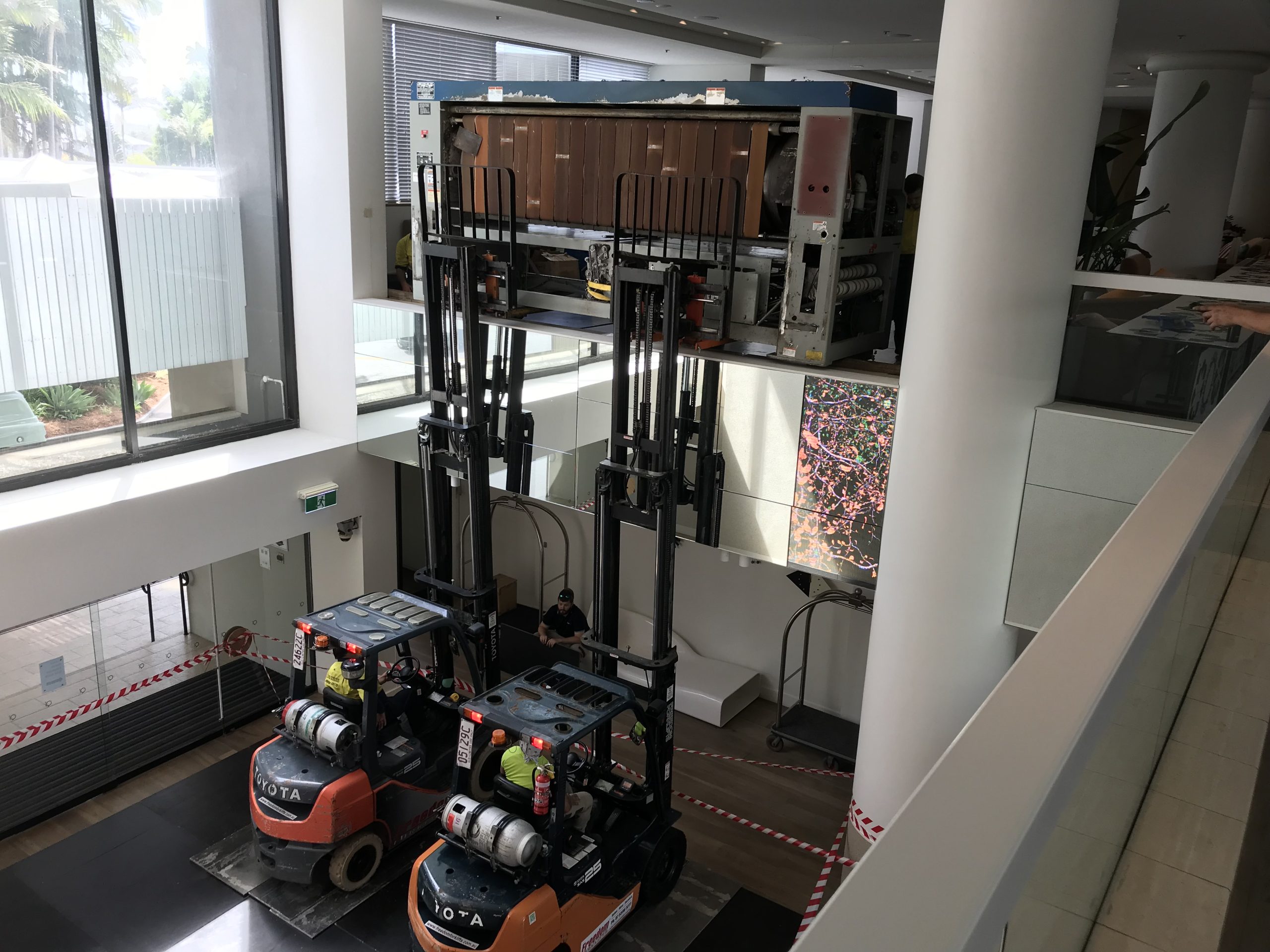 Industrial printer lifted with dual forklift onto elevated level.